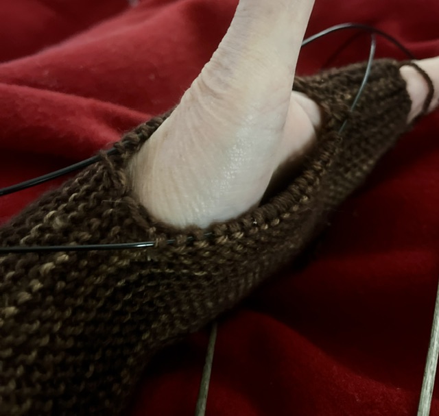 A knitted rectangle has been folded over, and the two ends have been fastened to each other with a seam that's open in the middle. A white person's hand is wearing the resulting tube with the thumb sticking out the opening in the seam. The stitches around the hole are still live stitches on knitting needles.